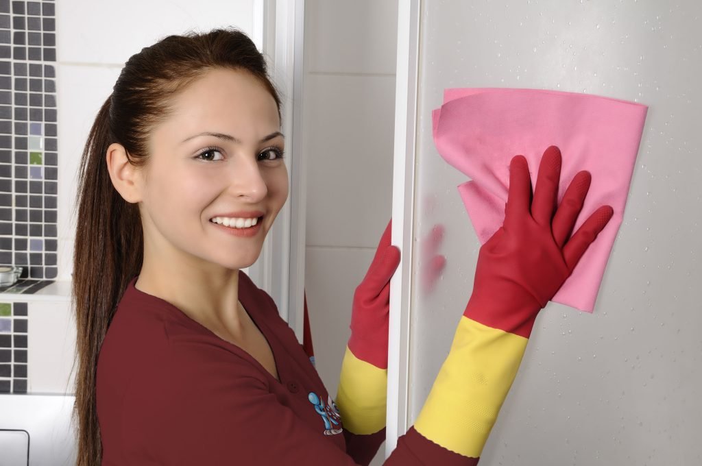 Woman cleaning a shower with pink cloth, wearing red and yellow rubber gloves, promoting cleaning services for allergy relief.