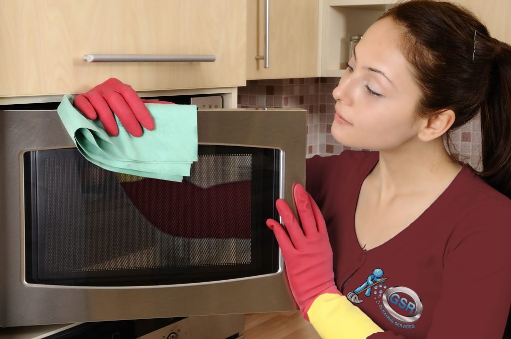 Woman in red and yellow gloves cleaning a microwave with a green cloth, promoting kitchen cleanliness and maintenance tips from GSR Cleaning Services.