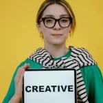 Woman in green top and striped scarf holding a sign that reads "CREATIVE," set against a bright yellow background, symbolizing innovative and professional cleaning services.