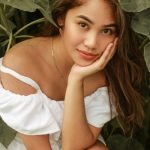 Young woman with long hair, resting her chin on her hand, wearing a white off-shoulder top, surrounded by lush green foliage, conveying a sense of relaxation and contentment.